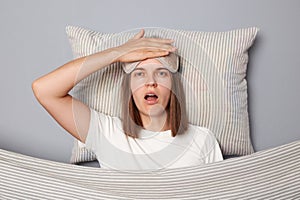 Shocked sad woman in white T-shirt and sleeping eye mask lie in bed on pillow under blanket isolated on gray background being