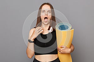 Shocked scared astonished girl holding fitness mat in her hands posing isolated over gray background looking at camera with open