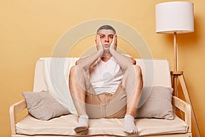 Shocked scared brunette young man wearing casual white t shirt sitting on sofa against beige wall keeps hands on cheeks looking at