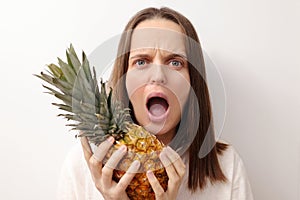 Shocked scared Caucasian woman with brown hair holding fresh ripe pineapple isolated over gray background screaming with negative