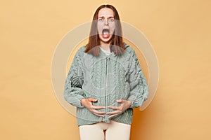 Shocked scared pregnant woman wearing knitted warm sweater standing isolated over beige background touching her belly screaming