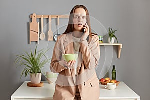 Shocked scared woman with brown hair wearing beige jacket standing in home kitchen interior talking on smart phone while having