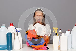 Shocked scared woman posing at workplace with cleaning detergents isolated over gray background holding clothing un ahnds screming