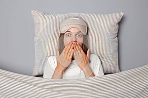 Shocked scared woman in white T-shirt and sleeping eye mask lie in bed on pillow under blanket isolated on gray background