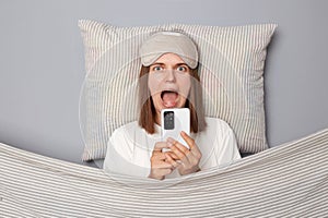 Shocked scared young woman wearing white T-shirt and sleep eye mask lies under blanket in bedroom isolated on gray background