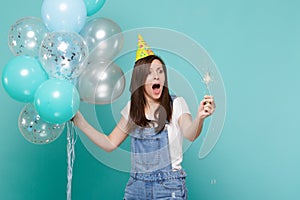 Shocked screaming woman in birthday hat looking on burning sparkler, celebrating, holding colorful air balloons isolated