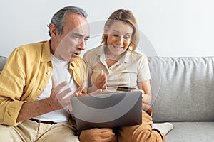 Shocked senior man looking at laptop screen and saw how much money his wife spend, woman holding credit card