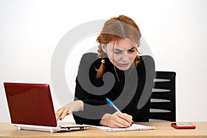 Shocked serious young office worker woman sitting behind working desk with laptop computer, cell phone and notebook