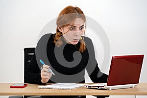 Shocked serious young office worker woman sitting behind working desk with laptop computer, cell phone and notebook