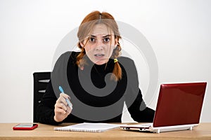 Shocked serious young office worker woman sitting behind working desk with laptop computer, cell phone and notebook