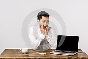 Shocked, speechless, astonished asian office guy sitting desk at work, reacting shook and surprised as looking laptop