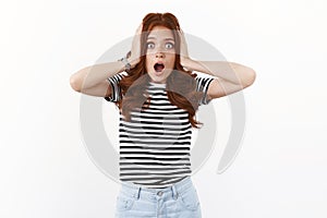 Shocked speechless nervous young cute redhead woman in striped t-shirt grab head in hands, open mouth gasping ambushed