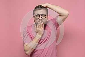 Shocked, strange man with glasses in fright covers mouth with one hand, put the other on head and looks with big eyes.