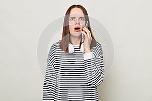 Shocked surprised brown haired adult woman wearing striped shirt standing isolated over gray background talking phone and looking