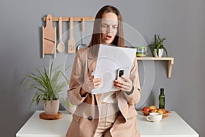 Shocked surprised disappointed woman with brown hair wearing beige jacket standing in home kitchen interior standing with smart