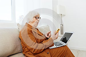 a shocked, surprised elderly woman is sitting in a bright apartment on a cozy sofa dressed in a brown suit and holding a