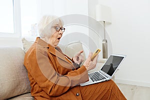 a shocked, surprised elderly woman is sitting in a bright apartment on a cozy sofa dressed in a brown suit and holding a