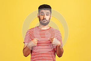 Shocked surprised man with beard in striped t-shirt pointing fingers at himself looking at camera with big eyes, astonishment