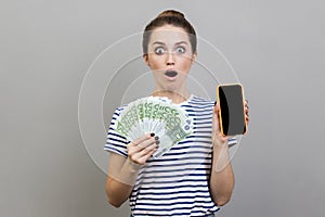 Shocked surprised woman holding fan of euro banknotes and smart phone with black blank screen.