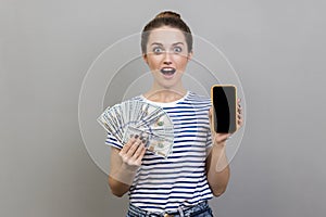 Shocked surprised woman holding in hands smart phone with blank screen and dollars banknotes.