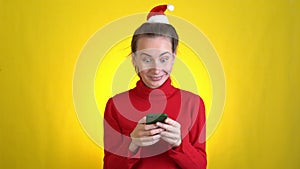 Shocked surprised woman Santa Christmas hat holding using mobile cell phone typing sms isolated on yellow background