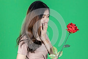 Shocked surprised young Asian woman holding rose over green isolated background. Love and Valentines day