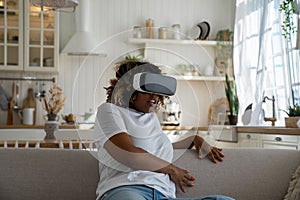 Shocked surprised young black woman sitting on sofa feeling scared in virtual reality