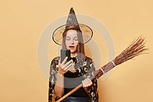Shocked surprised young woman wizard wearing witch costume holding in hand broom  over beige background holding mobile