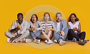 Shocked teenagers using phones, sitting on floor over yellow background