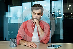 Shocked and upset young man sitting at the desk in the office, thoughtfully holding his head with his hand. Fired