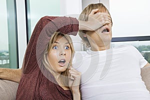 Shocked woman covering man's eyes while watching TV at home