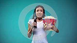 Shocked woman with cup of popcorn watching scary movie, holding fizzy water