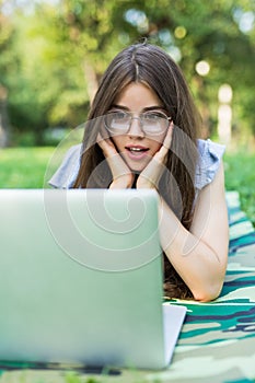 Shocked woman in eyeglasses lying on grass in park and using laptop computer