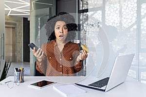 Shocked woman holding credit card and phone while working on laptop in modern office