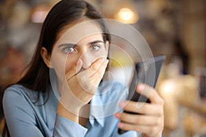 Shocked woman holding phone in a bar looks at you