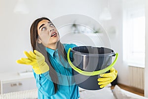 Shocked Woman Looks at the Ceiling While Collecting Water Which Leaks in the Living Room at Home. Worried Woman Holding Bucket