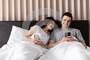 Shocked woman noticing her husband checking his smartphone and texting his lover while lying next to his wife in bed. Surprised