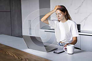 Shocked woman sitting in the kitchen and working on laptop and counting spendings and taxes