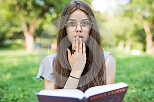 Shocked woman sitting in park while holding book and looking at the camera
