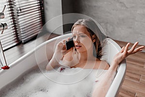 Shocked woman talking on cellphone while lying in bath, having conversation while relaxing in bathtub after hard day