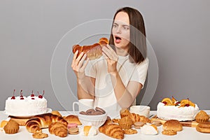 Shocked woman wearing white T-shirt sitting at festive table with various desserts, isolated over gray background holding baked