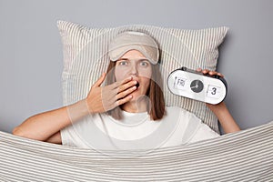 Shocked woman in white T-shirt and sleeping eye mask lie in bed on pillow under blanket isolated on gray background holding alarm