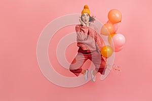 Shocked woman in yellow knitted hat jumping on pink backgorund. Studio shot of graceful girl with party balloons