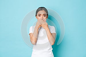 Shocked and worried asian woman in white t-shirt gasping, cover mouth and looking ambushed with bad concerning news