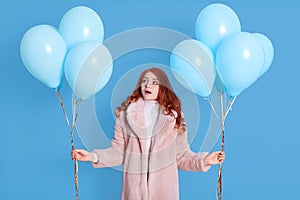 Shocked worried young ginger woman girl wearing fur coat, celebrating, posing on pastel color background with air balloons,