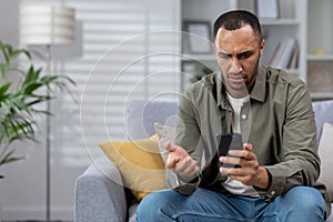 Shocked young african american man sitting on sofa at home with phone in hands and looking worriedly at screen. Lost