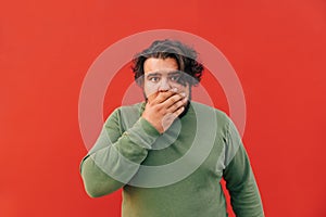Shocked young bearded guy is closing his mouth with his hand, standing on a red background with an astonished expression on his