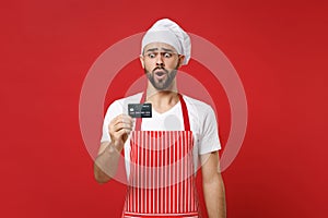 Shocked young bearded male chef cook or baker man in striped apron white t-shirt toque chefs hat isolated on red wall