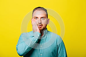 Shocked young bearded male with hand on face and looking at camera isolated on yellow