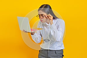 Shocked young business woman looking at laptop and holding glasses on yellow background